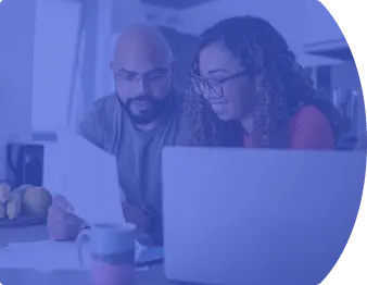 Um casal heterosexual sentado. Na mesa a frente dos dois há um notebook aberto, uma caneca e um celular. Os dois estão olhando para um papel que o homem está segurando