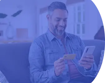 Homem sentado no sofá sorrindo, segurando o celular em uma das mãos e um cartão de crédito na outra