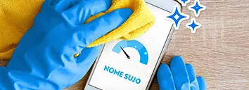 Celular em cima de uma mesa exibe na tela um ícone de score com o ponteiro bem baixo. Uma mão vestindo uma luva azul de limpeza passa um pano amarelo no celular. 